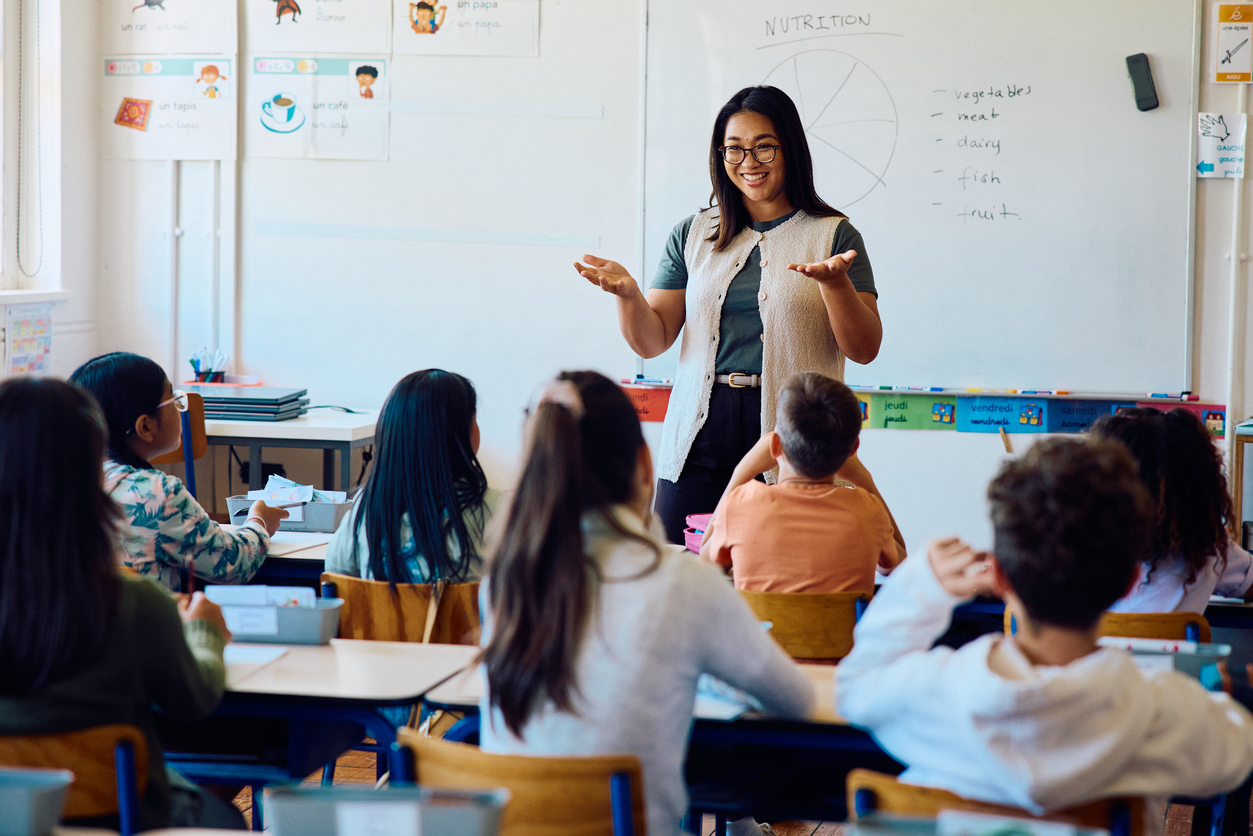 Happy elementary school teacher standing at the front of a classroom gesturing while explaining a lesson as students sit at desks and listen attentively.