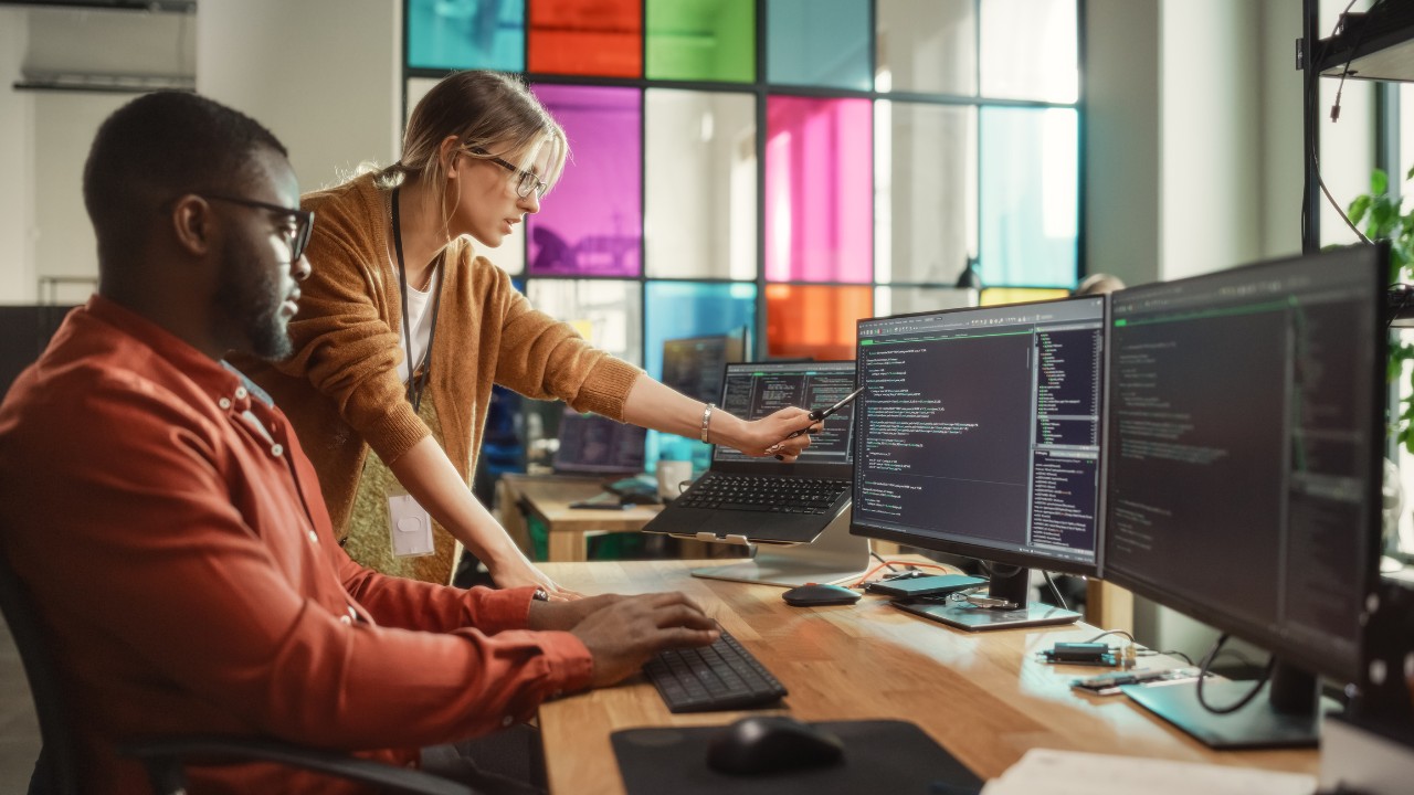 1481181765 African American Software Engineer Shows Code on Desktop Computer to Female Project Manager in Stylish Office. Black Man And Caucasian Woman Discussing Progress of AI Application Development.