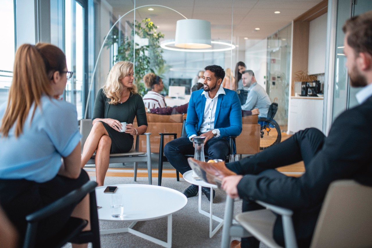 Diverse male and female businesspeople in conversation groups taking a break in modern office lobby.